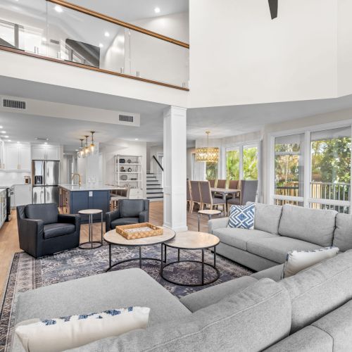 Gray sectional, two round tables, large windows, open kitchen and dining.