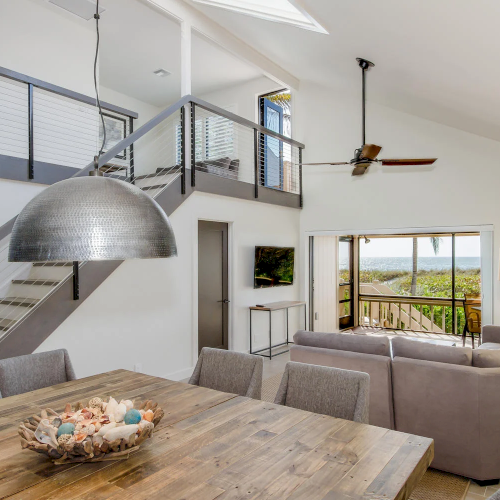 Wood table, gray chairs, sectional sofa, hanging light, stairs, ocean view.