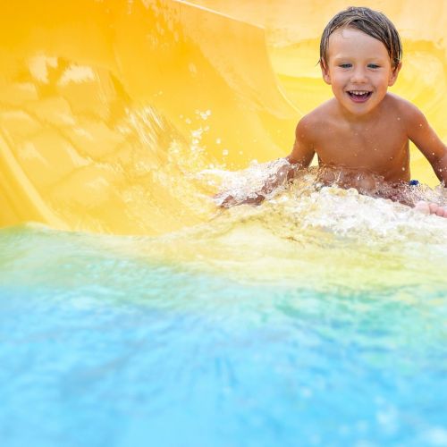 Cheerful blue-eyed kid on a water slide in the water park, a little boy merrily slides into the water along the slide in the amusement park, rest with children in the water park.