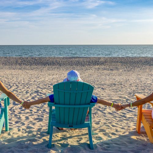 Three people sitting on colorful chairs facing the ocean, holding hands