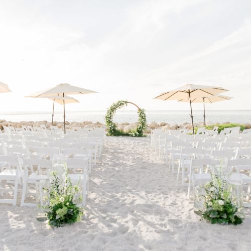 Rows of white chairs set up on the beach facing a round floral arch, with umbrellas in the background