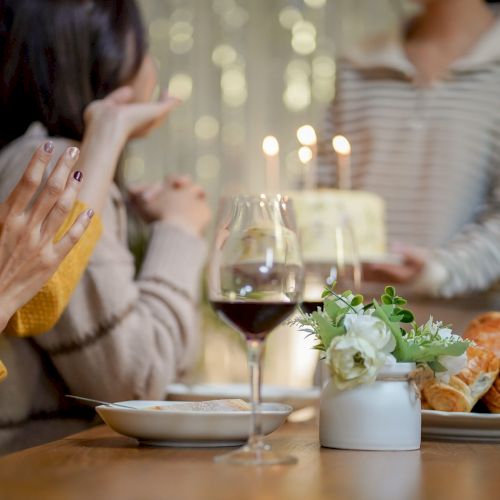 Cheerful friends enjoying home Birthday holiday party. Asian sister cheering drinking red wine celebrating with Birthday cake
