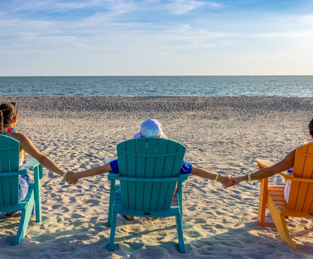 Three people sitting on colorful chairs facing the ocean, holding hands