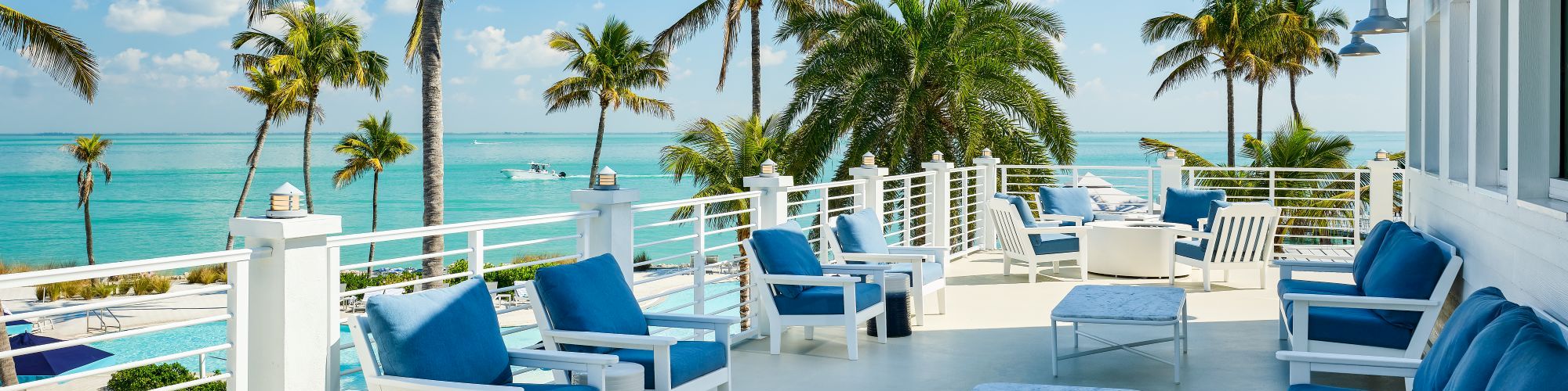 Oceanfront terrace with white railings, blue loungers, and tall palm trees under a bright blue sky.