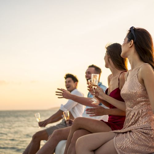 Group of diverse friends drink champagne while having a party in yacht. Attractive young men and women hanging out, celebrating holiday vacation trip while catamaran boat sailing during summer sunset.