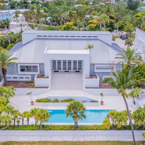 Aerial view of a white modern villa with a private blue pool, surrounded by palm trees and lush tropical landscaping.