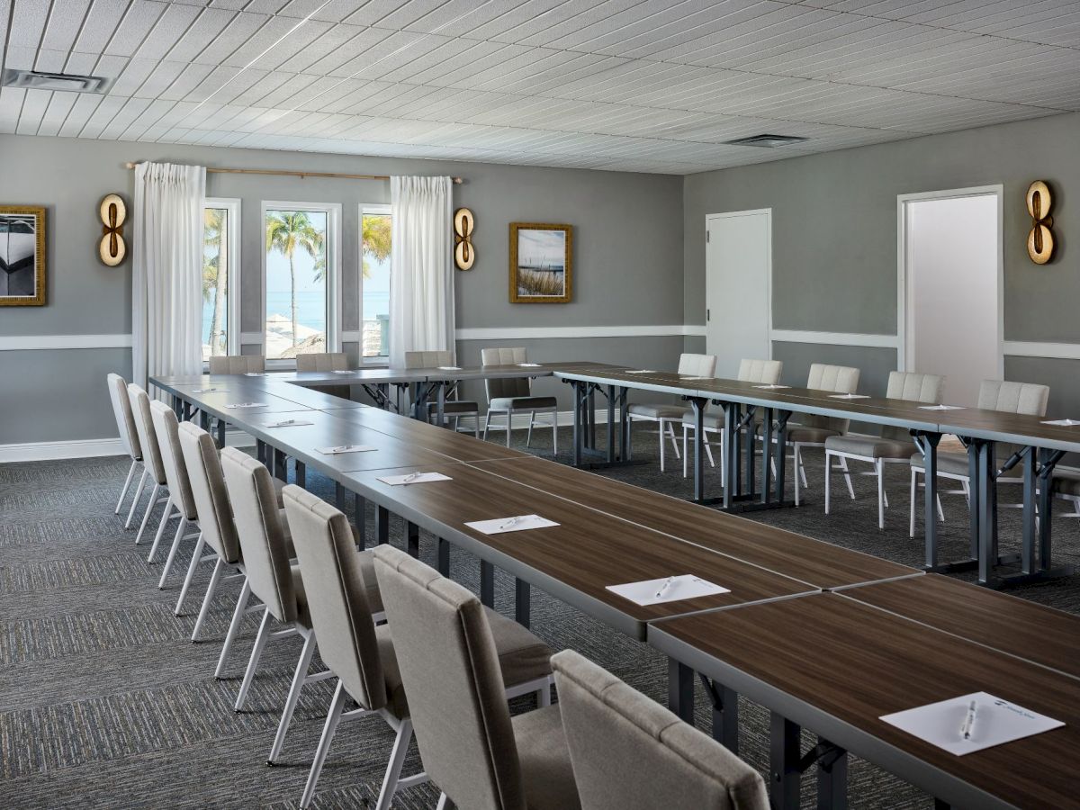 A modern conference room with a long wooden table, cushioned chairs, wall art, and ocean-view windows with white curtains.