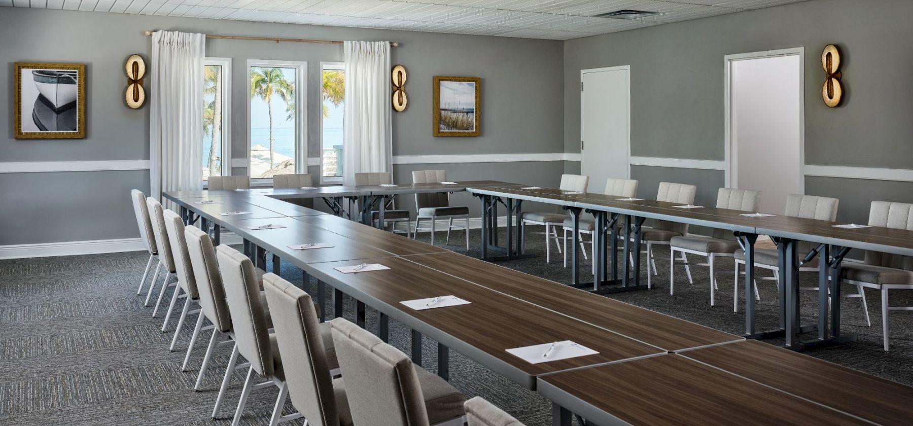 A modern conference room with a long wooden table, cushioned chairs, wall art, and ocean-view windows with white curtains.