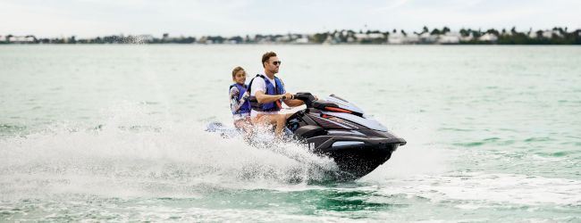 A man and a woman ride a jet ski across the ocean, splashing water on a sunny day.