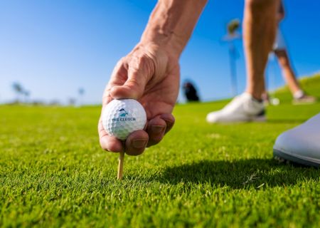 A golfer&rsquo;s hand holds a golf ball on a tee on a sunny green course, with another player and club in the background.