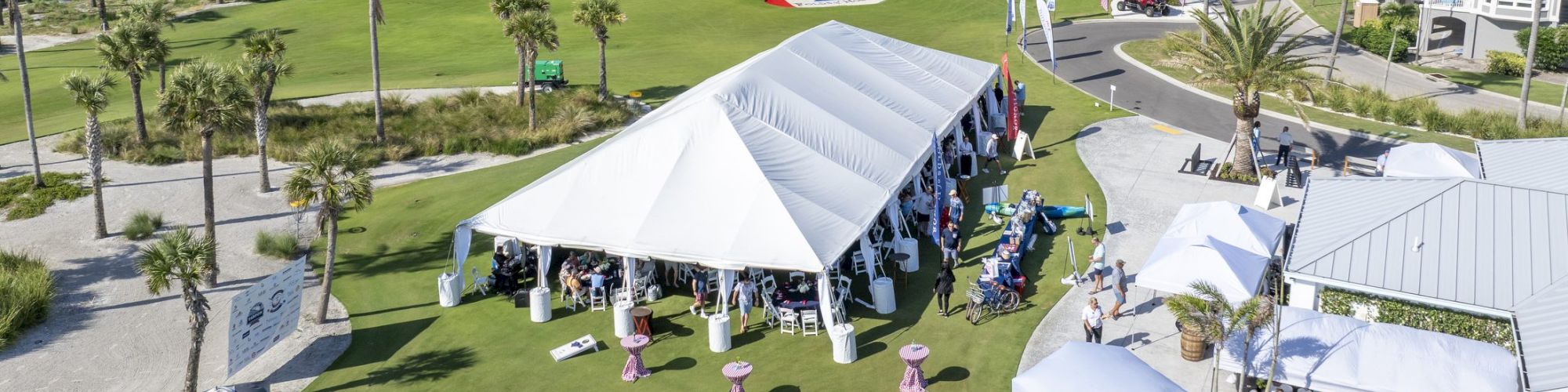 A sunny golf course by the ocean with white event tents, tables, chairs, and flags for a gathering.