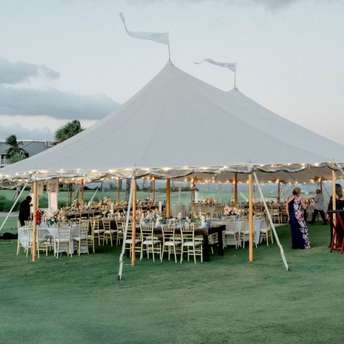 An outdoor event on a grassy field with a large white marquee tent, string lights, chairs, and tables for a celebration.