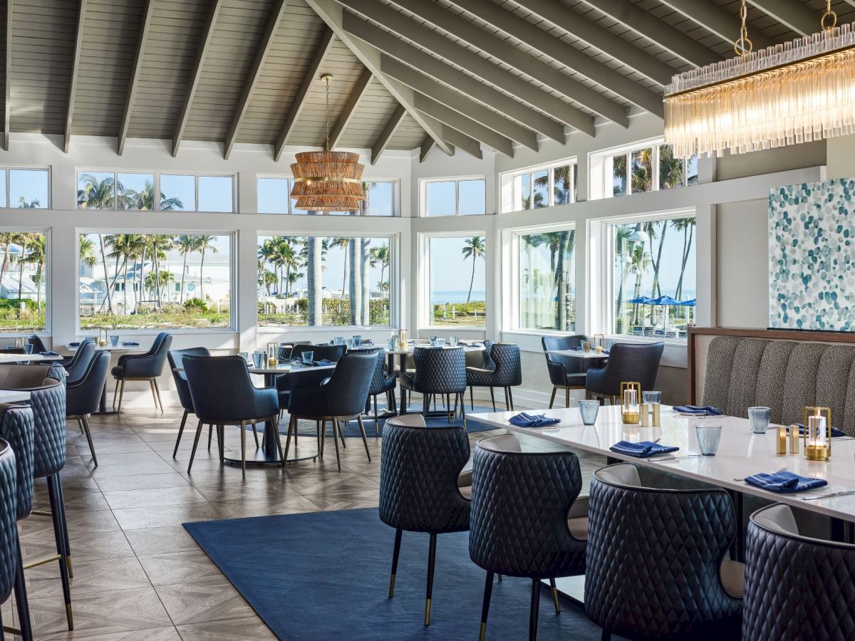 Bright seaside restaurant dining room with blue chairs, set tables, large windows with palm views, and a sparkling chandelier overhead.