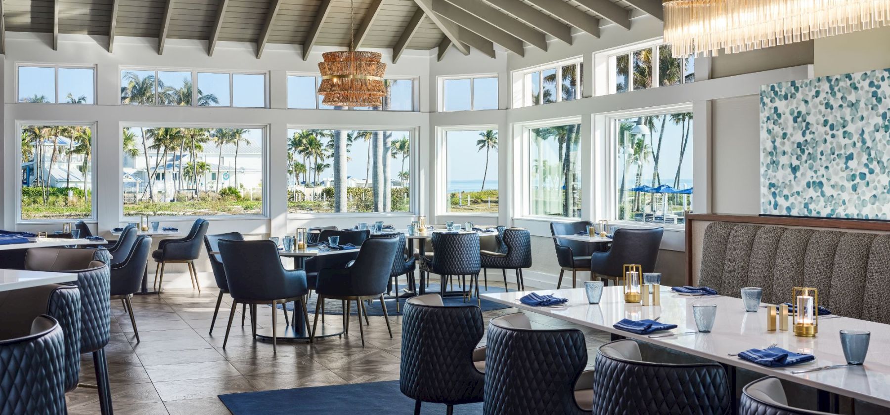 Bright seaside restaurant dining room with blue chairs, set tables, large windows with palm views, and a sparkling chandelier overhead.