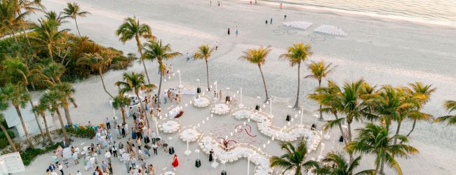 A beach wedding on the sand with palm trees, white chairs and round tables, guests gathered, and decorative candles.
