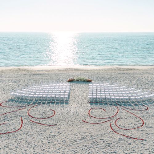 A beach wedding setup with chairs and flowers, decorative heart/letter designs in the sand, and the ocean horizon in the background.
