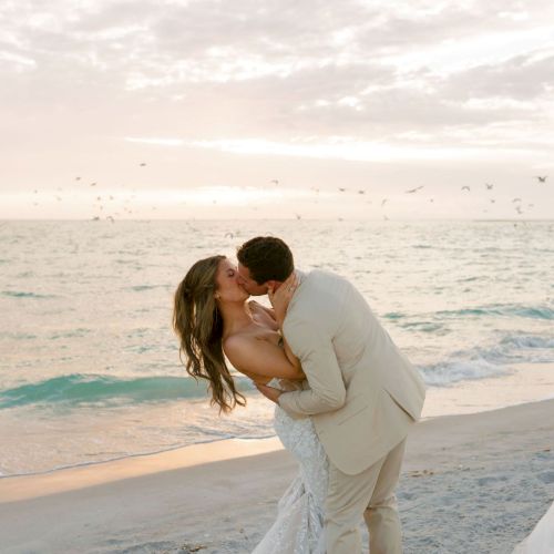 A bride and groom kiss on a sandy beach at sunset, ocean waves behind them and birds flying overhead, captured in a dreamy wedding photo.