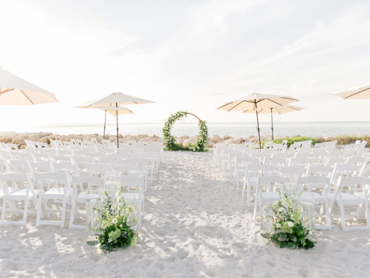 Rows of white chairs set up on the beach facing a round floral arch, with umbrellas in the background