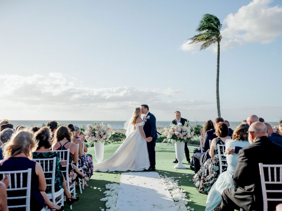 Outdoor beach wedding ceremony with a couple kissing under a tall palm tree, guests seated on chairs.