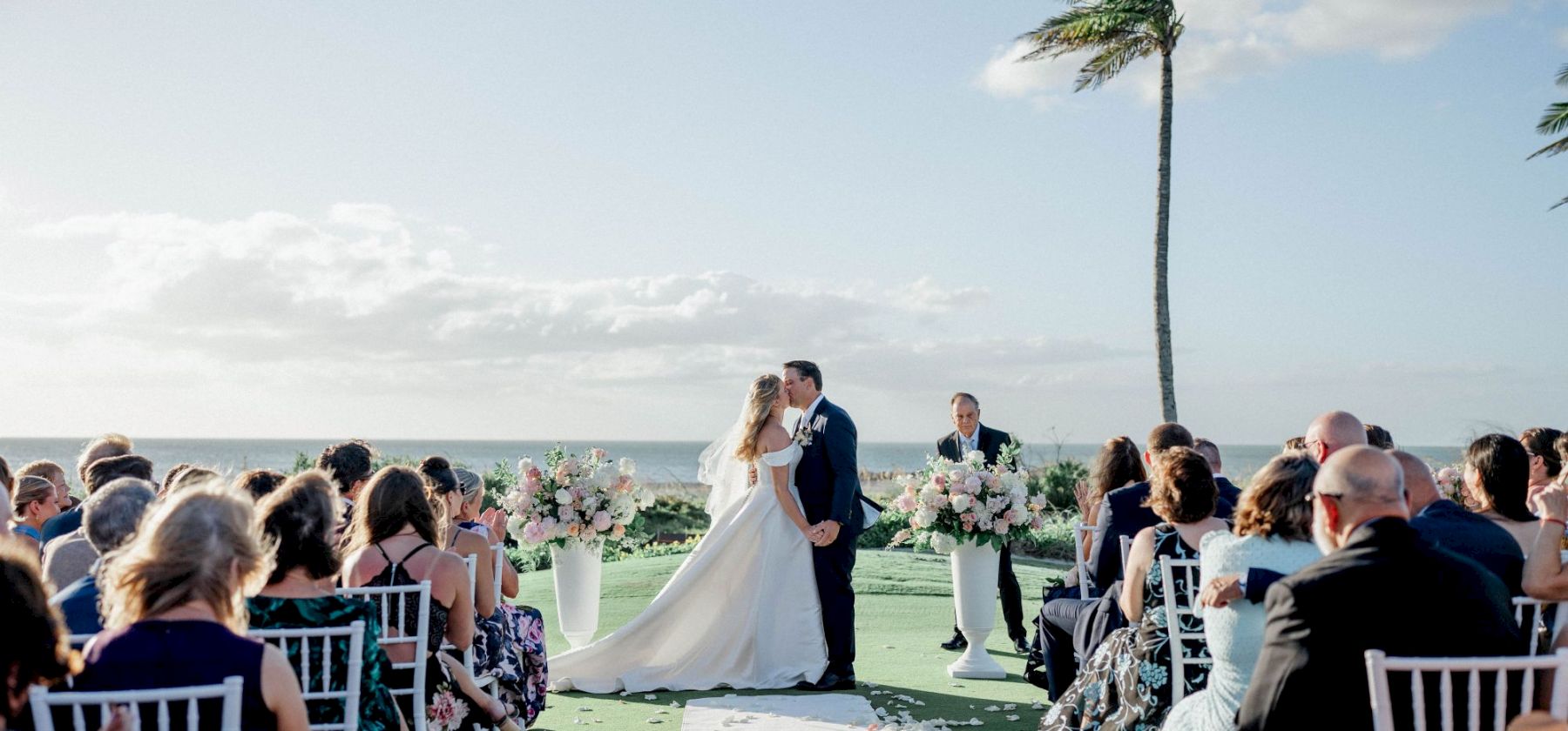 Outdoor beach wedding ceremony with a couple kissing under a tall palm tree, guests seated on chairs.