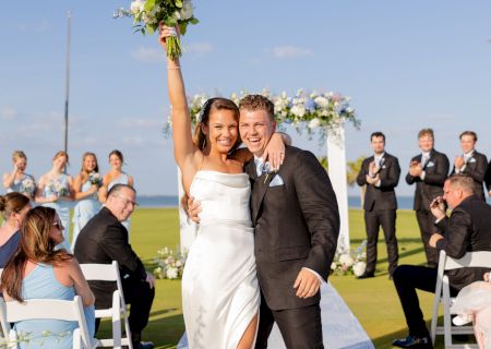 A wedding couple celebrates on a sunny outdoor lawn as the bride lifts a bouquet and guests clap in the background.