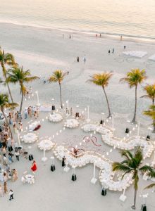 A beach wedding reception setup with guests, white tables, chairs, lanterns, and palm trees on a sandy shore at sunset.