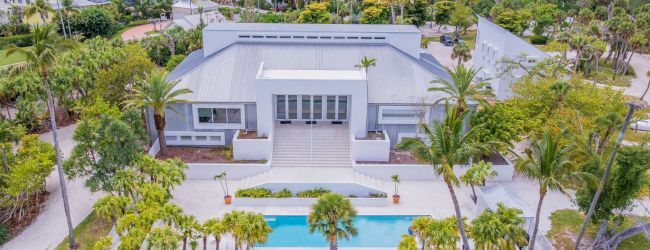 Aerial view of a modern white villa in a tropical neighborhood, surrounded by palms, with a bright blue swimming pool and gardens.