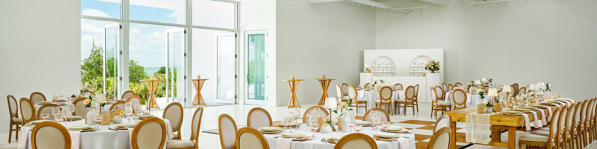 Bright event hall set for a wedding with banquet tables, wooden chairs, place settings, flowers, and large floor-to-ceiling windows.