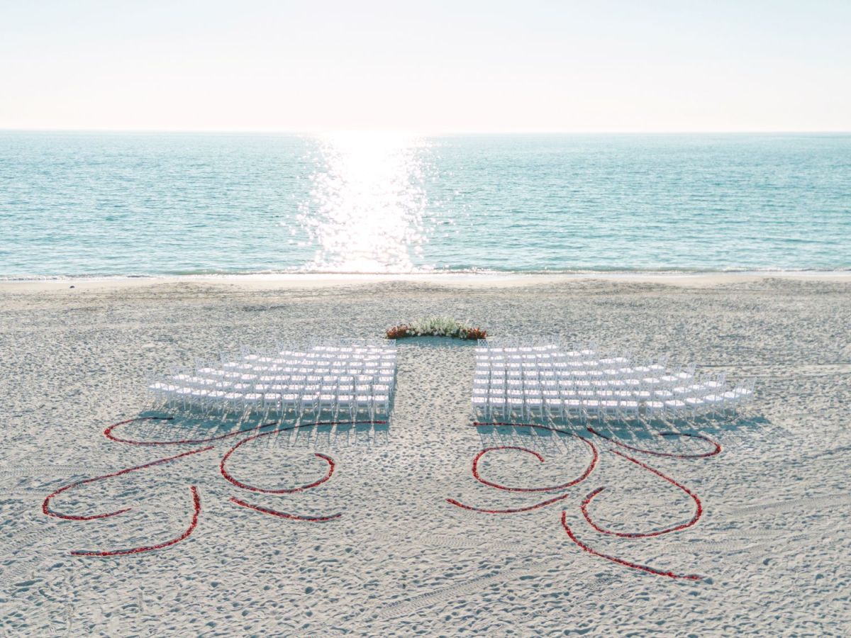 A sandy beach wedding setup with white chairs, red heart outlines, and a flower centerpiece facing the ocean.