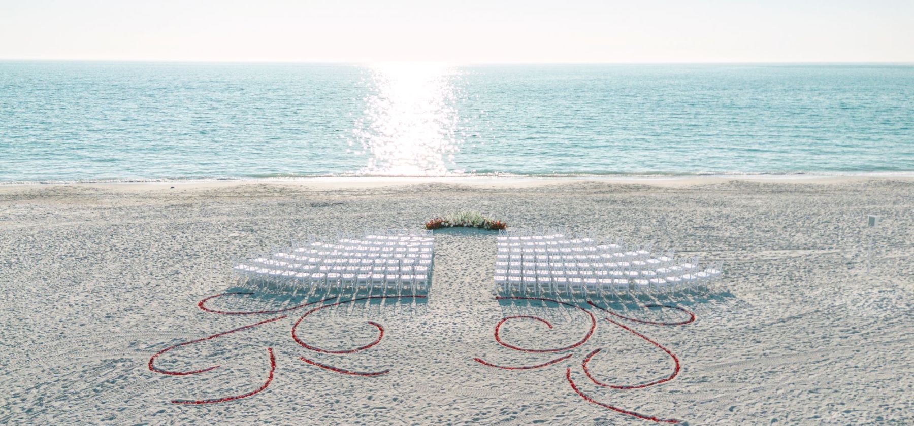 A sandy beach wedding setup with white chairs, red heart outlines, and a flower centerpiece facing the ocean.