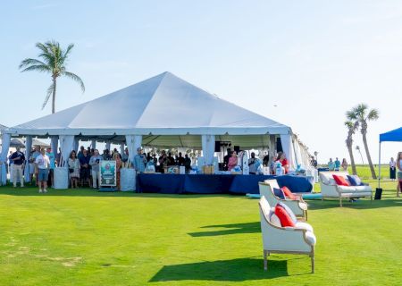 Outdoor charity event on a sunny green lawn with a large white tent, stage setup, and people gathered around nearby.