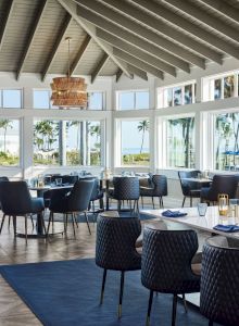 Bright modern dining room with blue chairs and set tables, a hanging crystal-like chandelier, and large windows showing palm-lined ocean views.