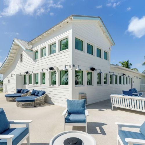 Sunny terrace beside a white house with blue lounge chairs, palm trees, and a clear sky.