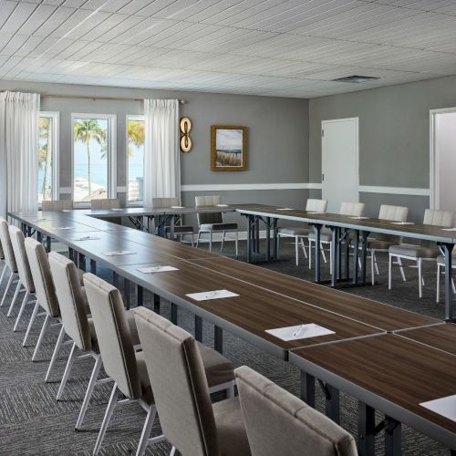 A modern conference room with a long meeting table, cushioned chairs, framed art, curtains, and windows showing palm trees outside.