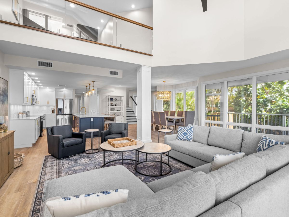 Gray sectional, two round tables, large windows, open kitchen and dining.