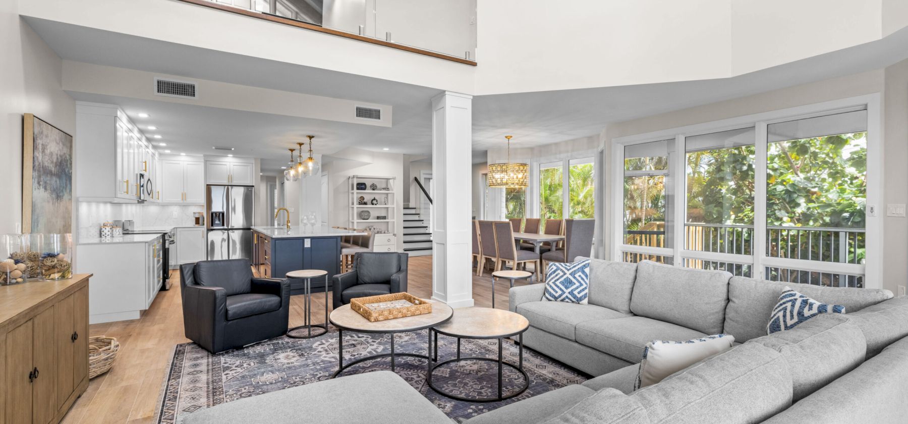 Gray sectional, two round tables, large windows, open kitchen and dining.