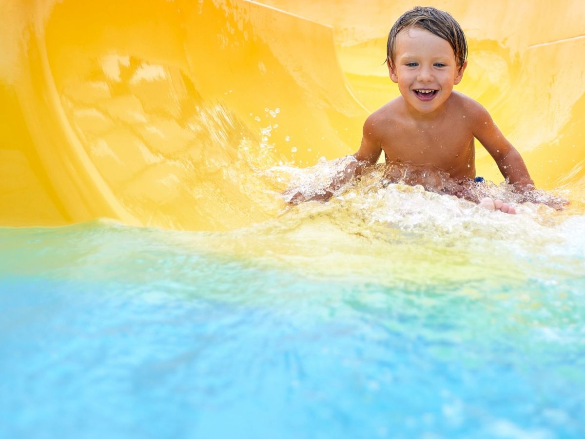 Cheerful blue-eyed kid on a water slide in the water park, a little boy merrily slides into the water along the slide in the amusement park, rest with children in the water park.