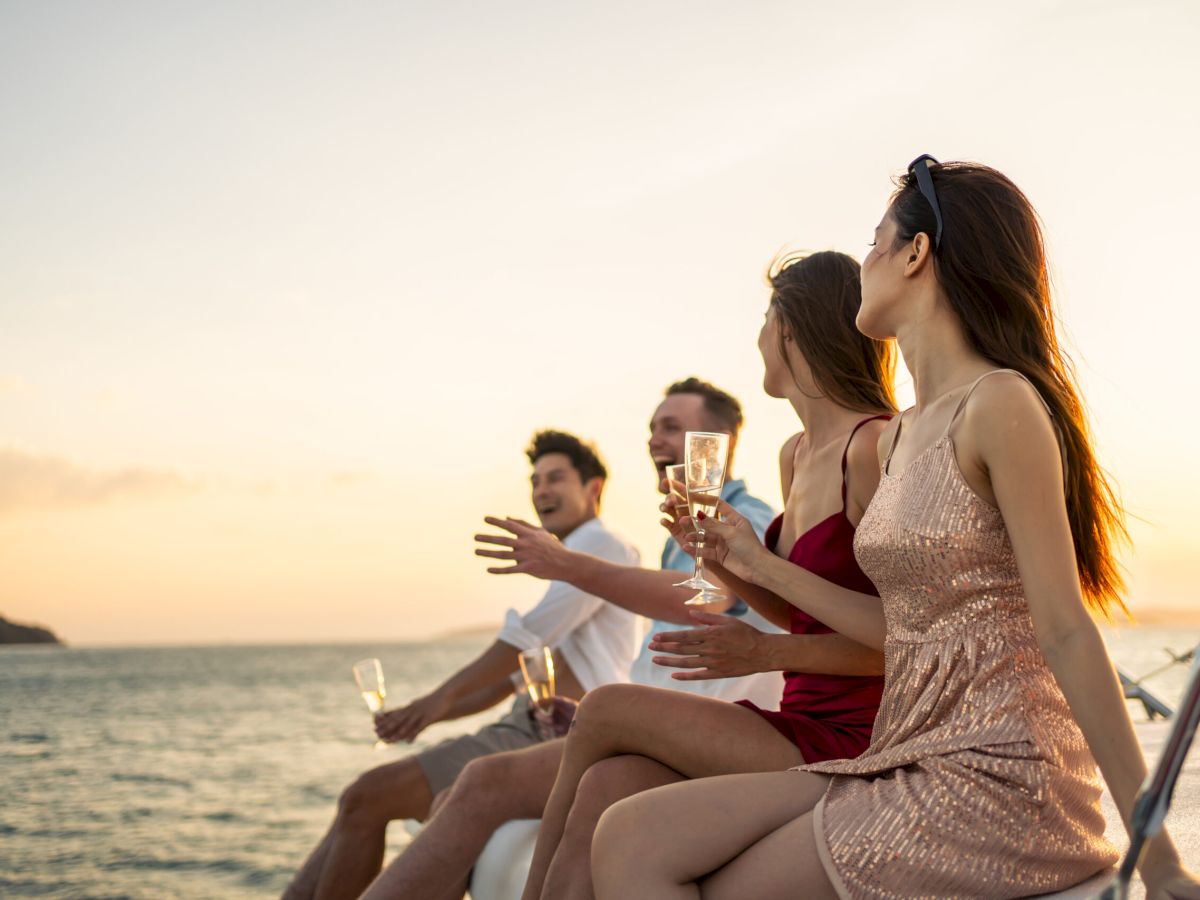 Group of diverse friends drink champagne while having a party in yacht. Attractive young men and women hanging out, celebrating holiday vacation trip while catamaran boat sailing during summer sunset.