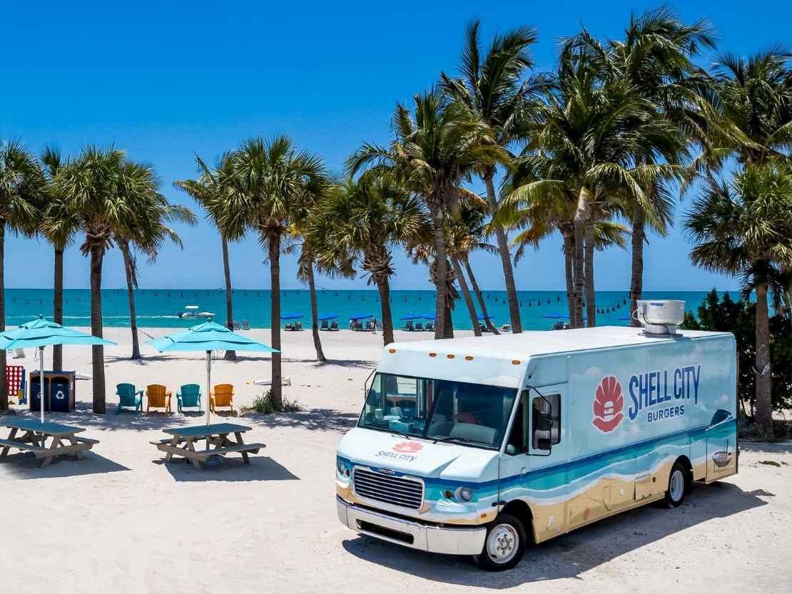 Shell City truck parked on the beach with palm trees, picnic tables, and ocean in the background