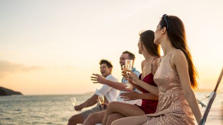 Group of diverse friends drink champagne while having a party in yacht. Attractive young men and women hanging out, celebrating holiday vacation trip while catamaran boat sailing during summer sunset.