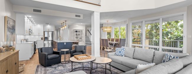 A bright modern living room with gray sofas, a coffee table, and a view to tropical greenery, opening into a sleek kitchen.