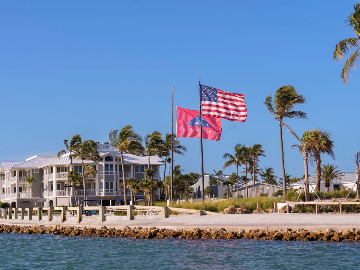 Two American flags fly in front of palm trees and seaside buildings under a clear blue sky.