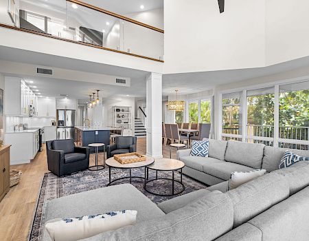 Gray sectional, two round tables, large windows, open kitchen and dining.