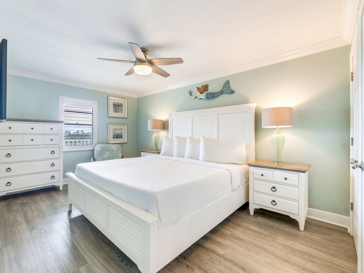 Bright and airy bedroom with a white king-size bed, matching furniture, coastal wall art, and wood-style flooring.