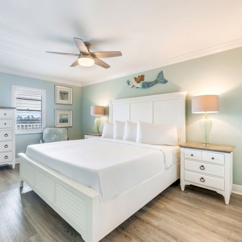 Bright and airy bedroom with a white king-size bed, matching furniture, coastal wall art, and wood-style flooring.