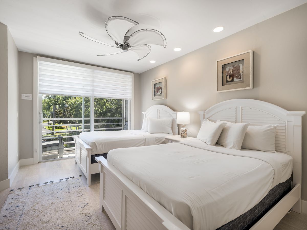Bright bedroom with two white beds, large window, and modern ceiling fan
