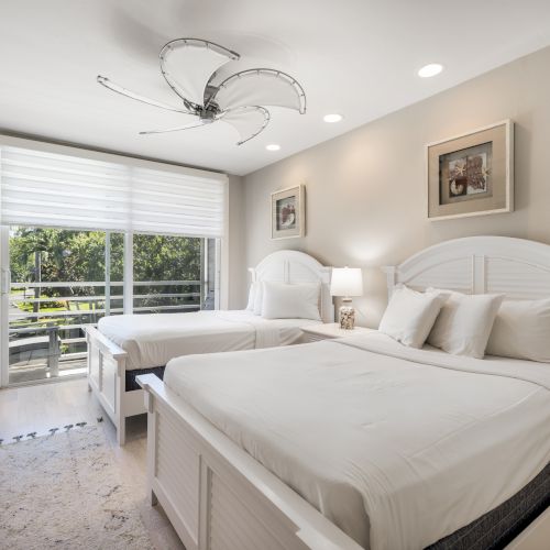 Bright bedroom with two white beds, large window, and modern ceiling fan
