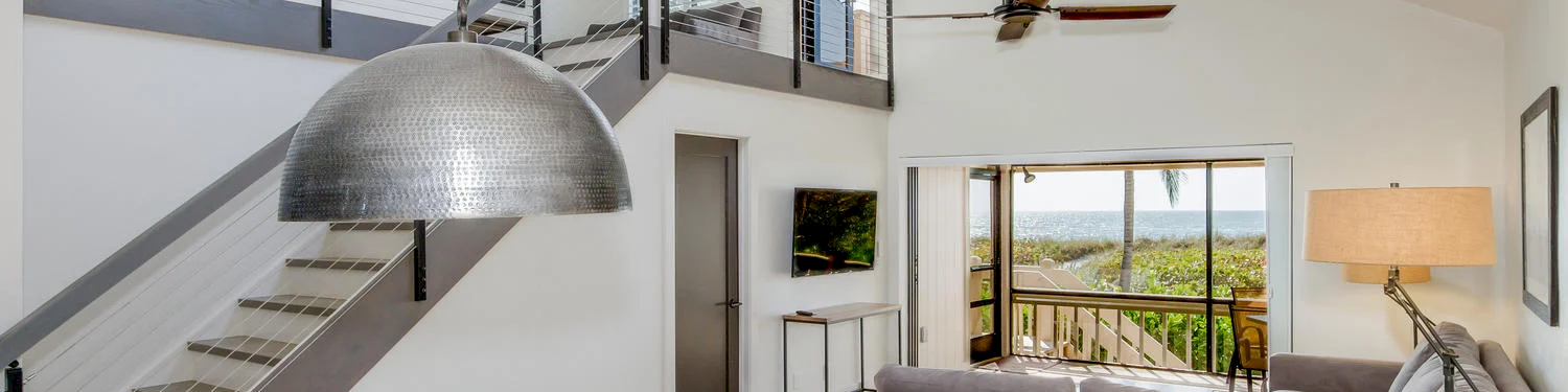 Wood table, gray chairs, sectional sofa, hanging light, stairs, ocean view.