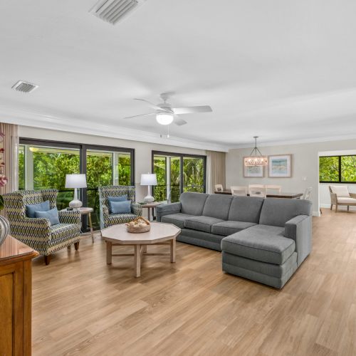 Living room with a gray sectional sofa, two armchairs, a round coffee table, and large windows showing greenery