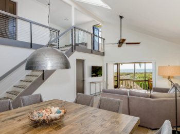 Wood table, gray chairs, sectional sofa, hanging light, stairs, ocean view.